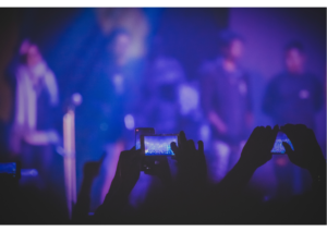 Ein Bild von einem Konzert aus der Perspektive des Publikums. Man sieht Hände von den Konzertgängern, die Bilder und Videos von der Band auf der Bühne machen.